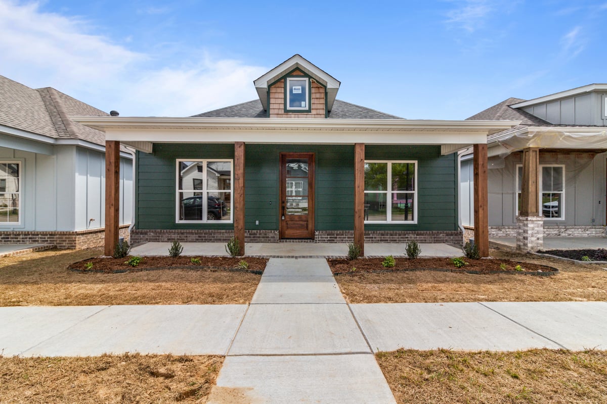 Single-story house with a green exterior, wood columns, a covered front porch, and a glass front door. The yard has a concrete walkway and young landscaping, with neighboring houses visible on both sides.