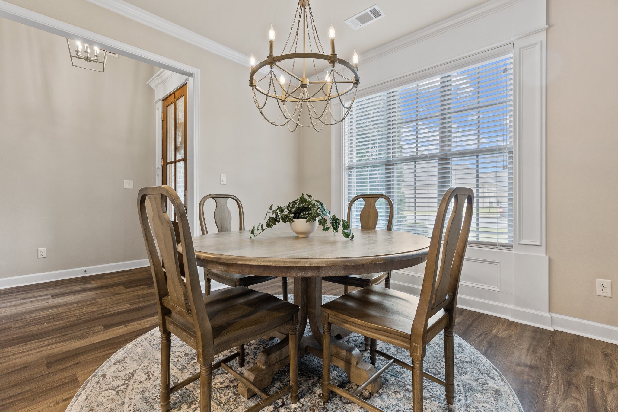 A dining room with a wooden round table, four wooden chairs, a decorative plant centerpiece, a chandelier above, large windows with blinds, wood flooring, and neutral-colored walls.