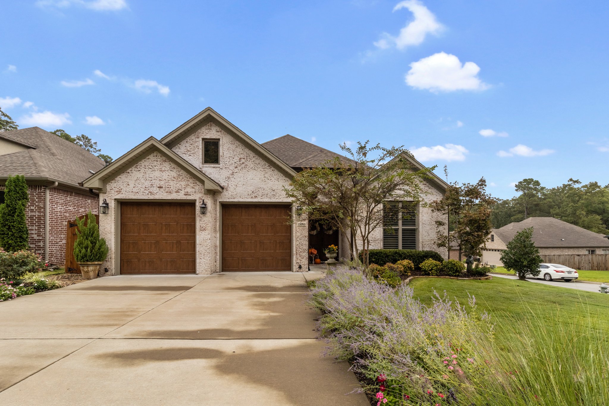 A modern suburban house with two wooden garage doors, a light brick exterior, and a driveway bordered by green grass and lavender plants under a blue sky with scattered clouds.