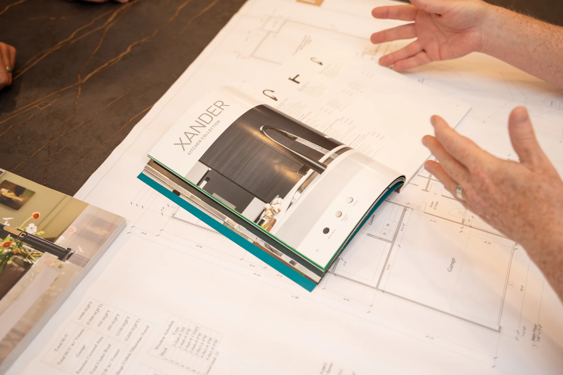 A person gestures with their hands over a table covered with architectural plans and an open catalog displaying a modern kitchen design labeled XANDER.