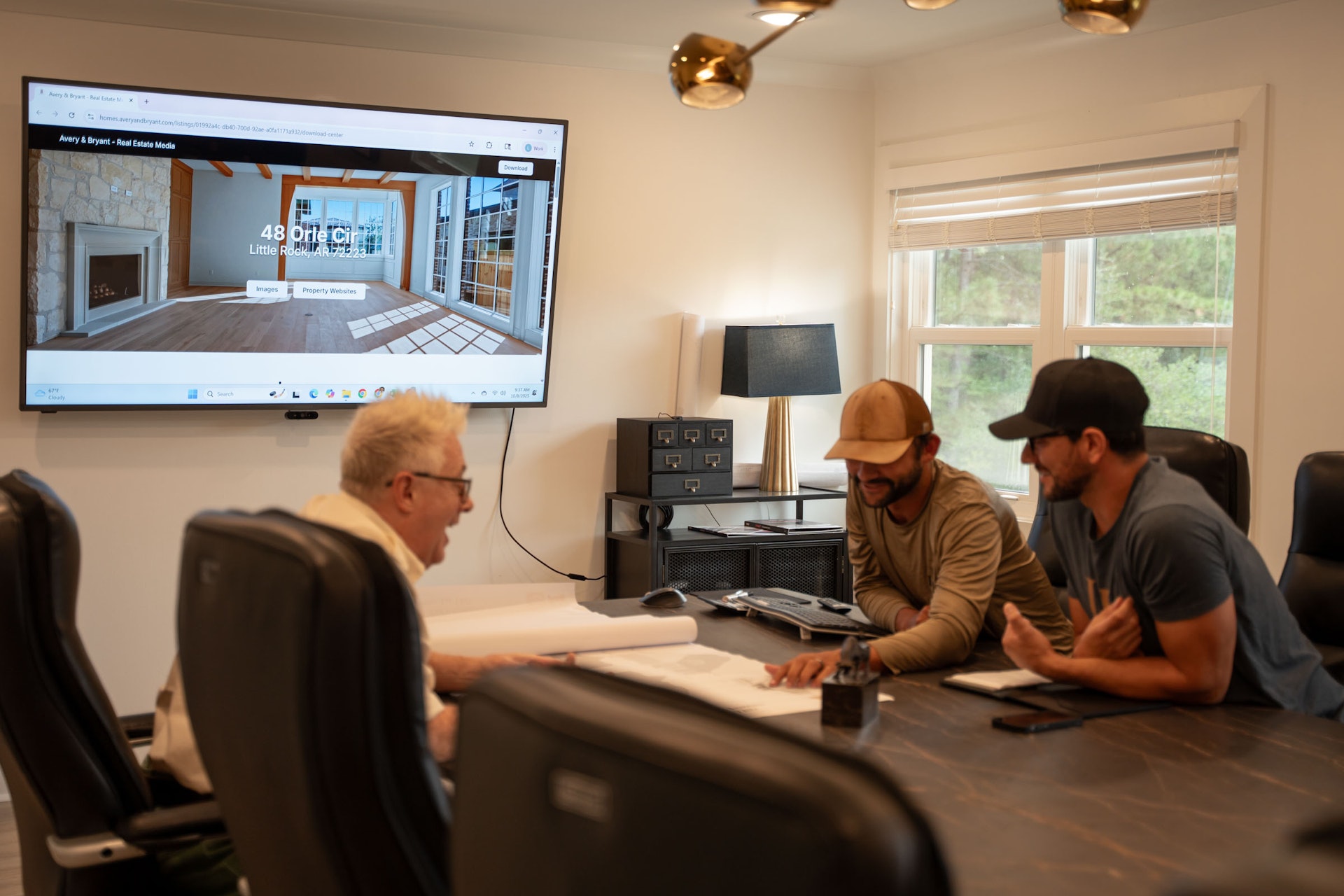 Three men sit at a conference table reviewing documents, while a large screen on the wall displays a real estate listing for 48 Drift Cliff. The room has modern decor, with a lamp and windows bringing in natural light.