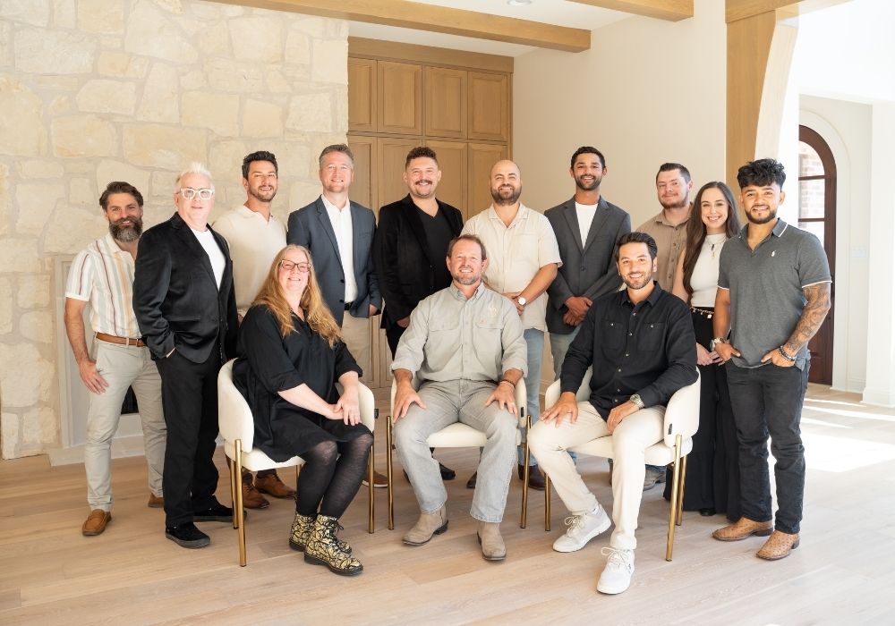 A group of fourteen people, dressed in business casual attire, pose and smile together indoors. Three people are seated in front, while the rest stand around them in a bright, modern room with wooden and stone accents.
