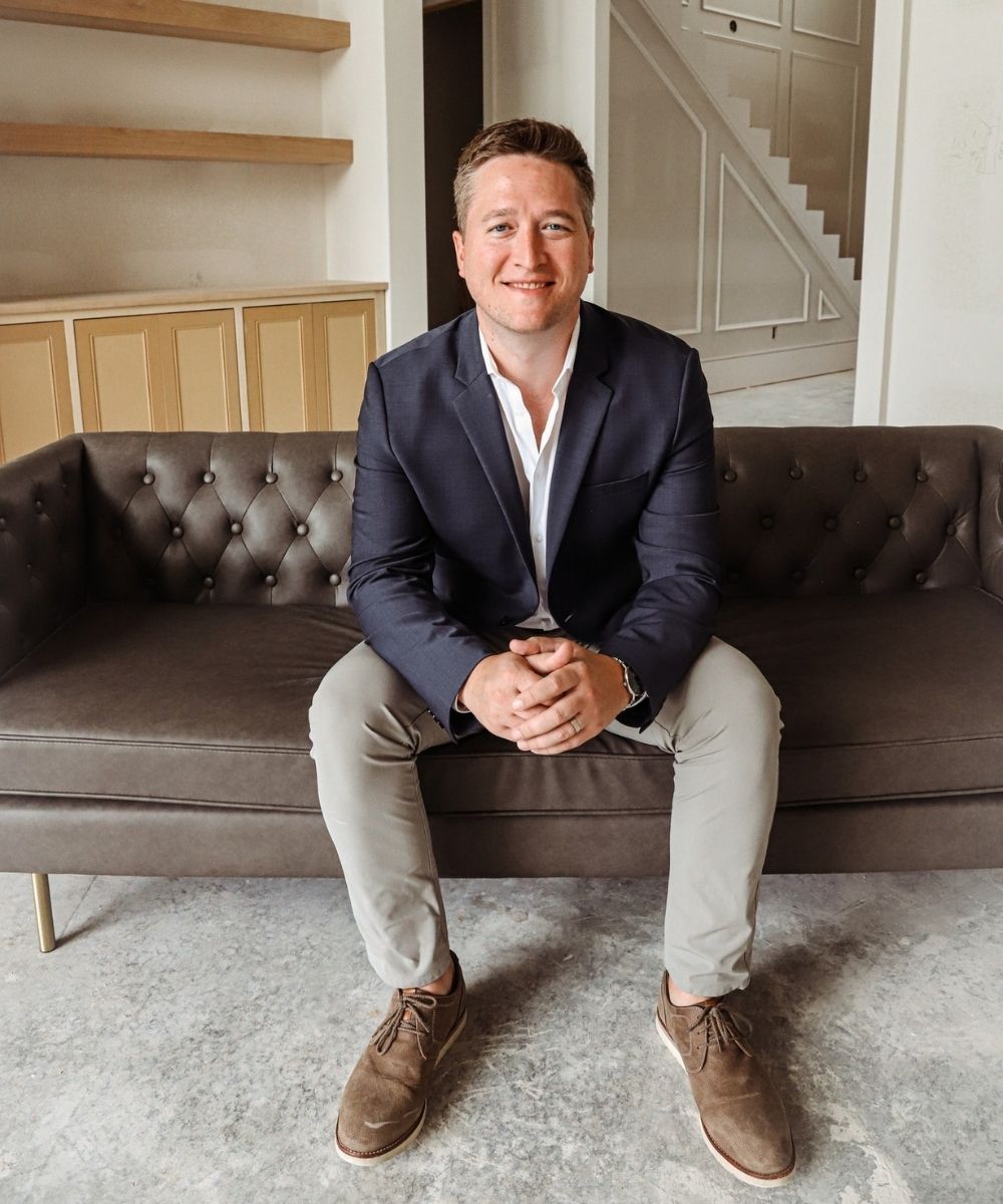 Jonathan Stone in a navy blazer and light pants sits smiling on a dark tufted sofa in a modern, unfinished room with shelves and stairs visible in the background.