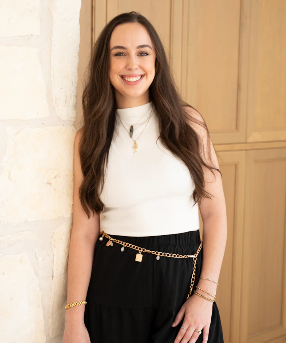 Lawson Maddox Richards with long brown hair stands smiling against a light stone wall. She wears a sleeveless white top, a black skirt with a gold chain belt, and layered gold necklaces and bracelets. Light wooden cabinets are in the background.