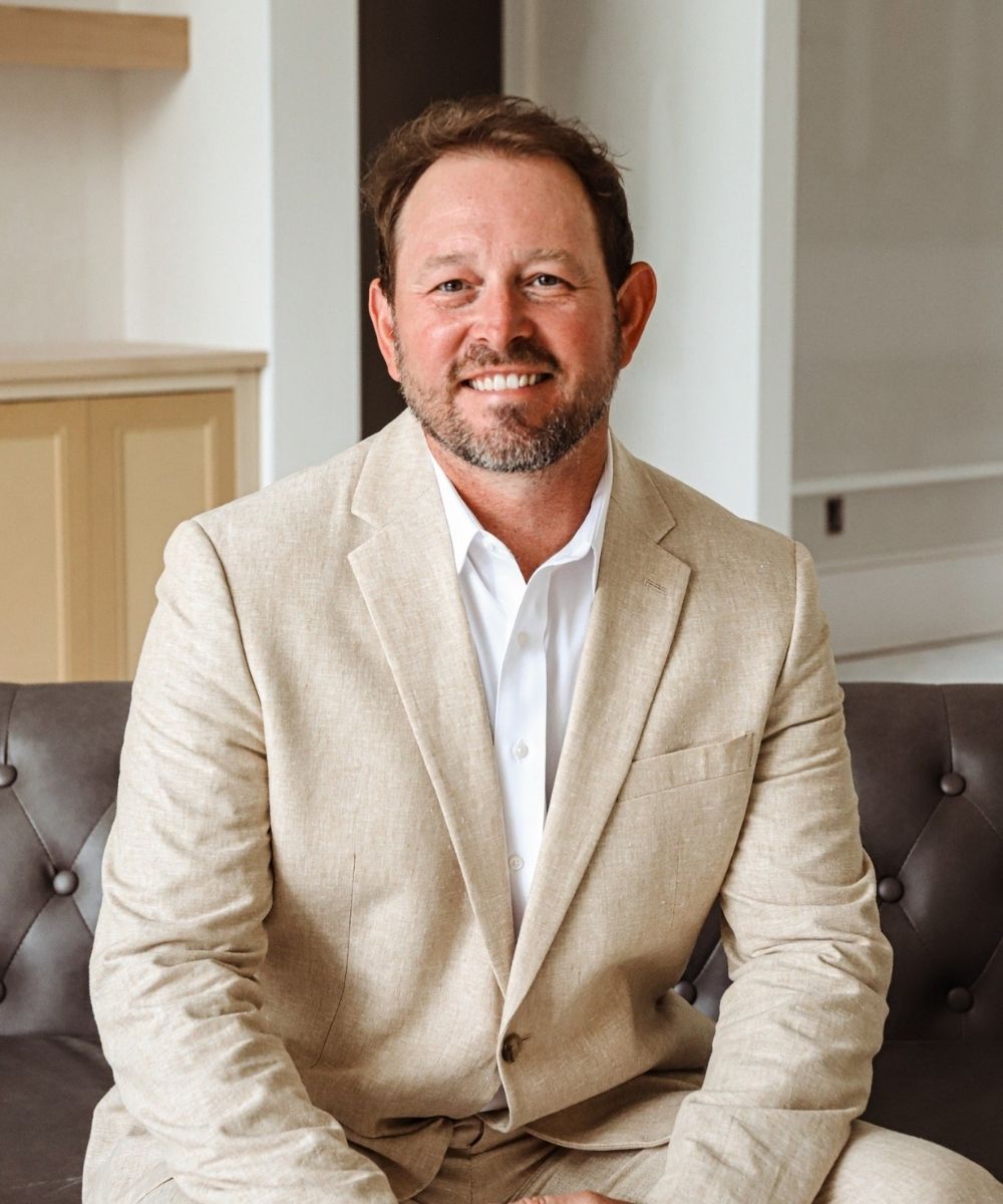 Scott Hurley with short brown hair and a beard, wearing a beige suit and white shirt, sits on a dark tufted sofa, smiling in a modern, light-filled room.