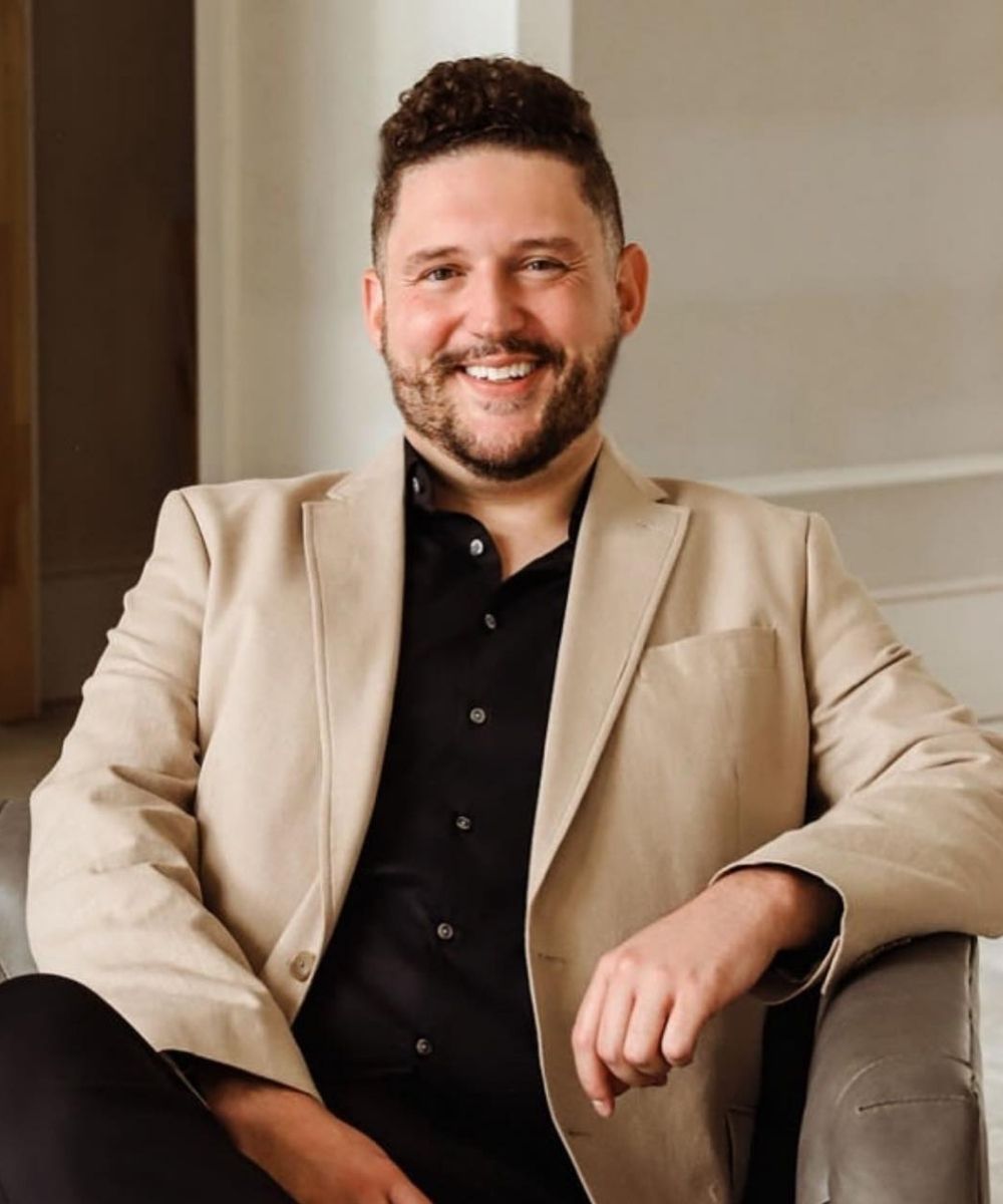 Trey Clifton with short curly hair and a beard, wearing a beige blazer over a black shirt, sits and smiles at the camera in a light-colored room.