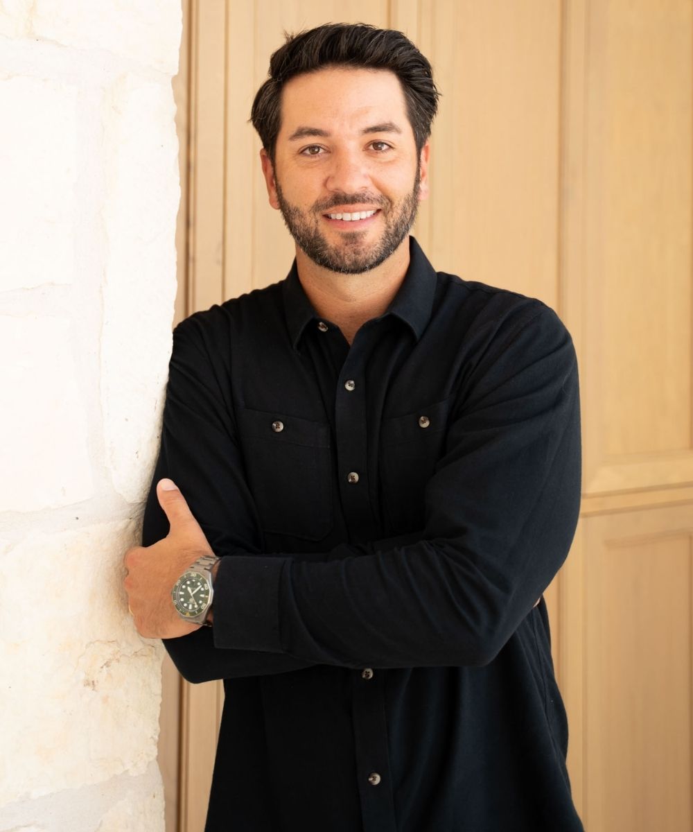 Zack DeYmaz with dark hair and a beard, wearing a black button-up shirt and a wristwatch, leans against a light-colored wall and smiles at the camera. Light wood paneling is in the background.