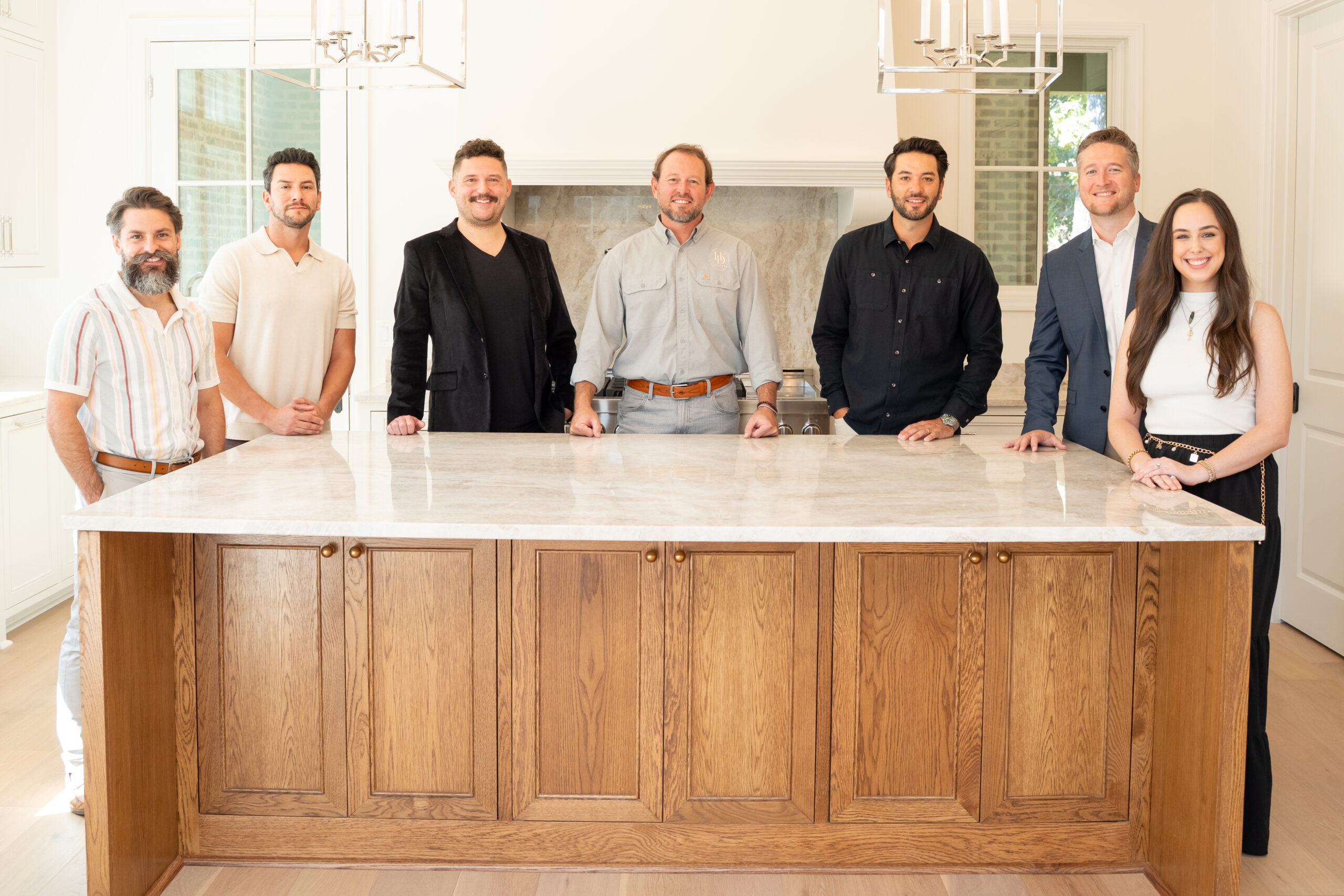 Seven people stand behind a large wooden kitchen island, smiling at the camera in a bright, modern kitchen with light walls and hanging lights above.