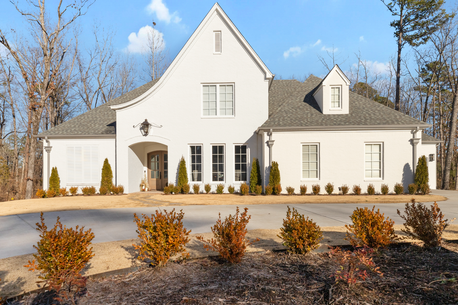 A large, modern white house with steep gray roof, multiple windows, and trimmed bushes, sits on a wide concrete driveway with a landscaped front yard and leafless trees in the background.