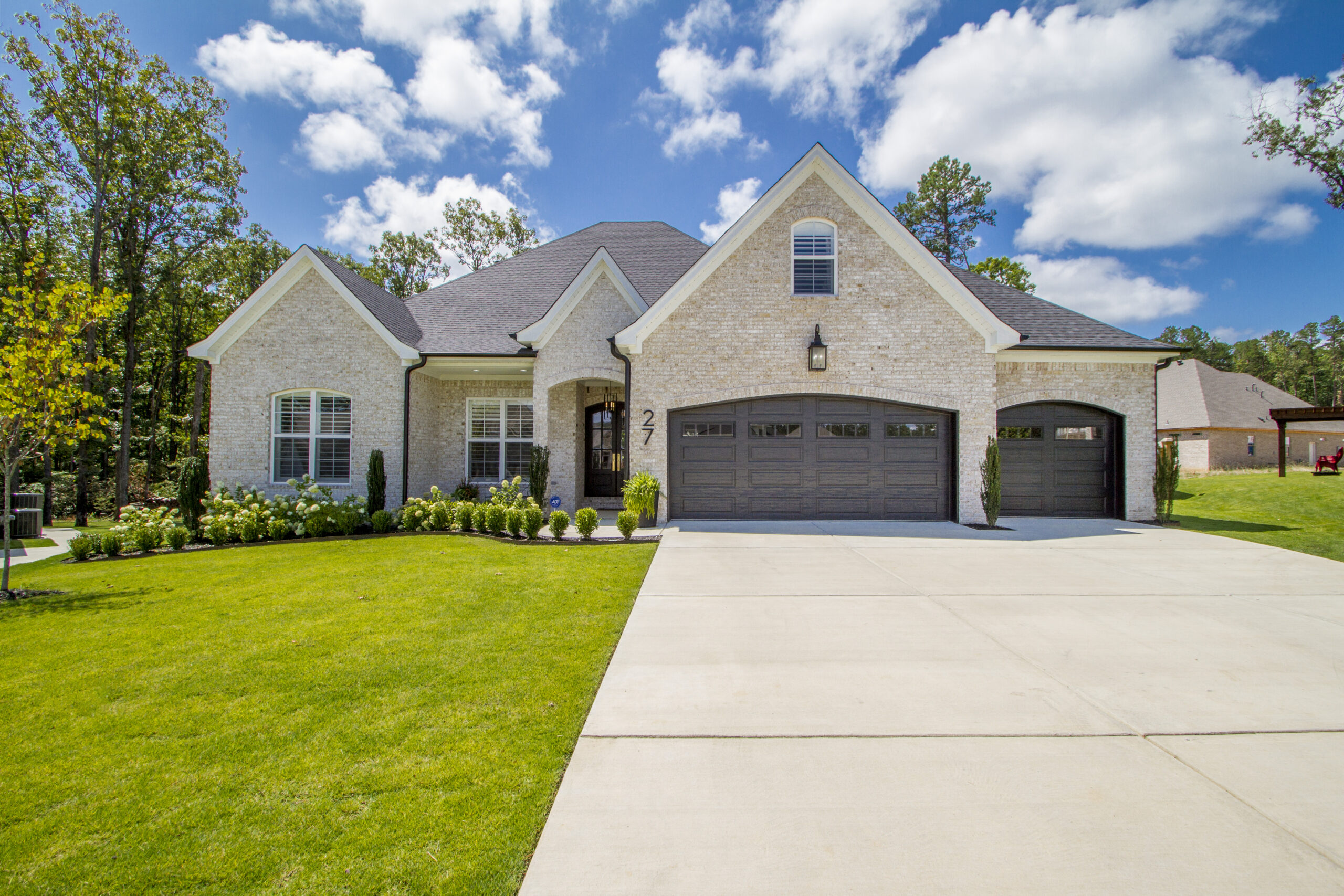 A modern, light brick house with dark wooden double garage doors, a large driveway, manicured lawn, and green landscaping, set under a bright blue sky with scattered clouds and trees in the background.