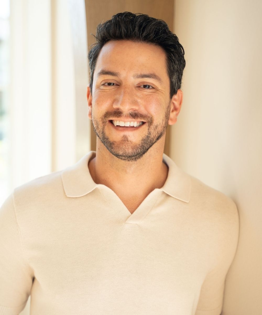 A man with short dark hair and a trimmed beard smiles warmly while standing indoors. He is wearing a light beige collared shirt and is positioned against a softly lit, neutral-colored wall.