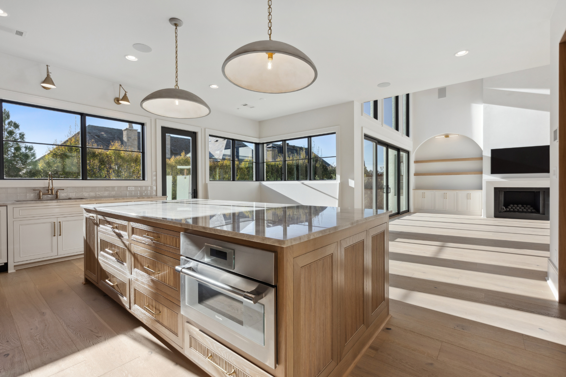 Modern kitchen with large marble island, built-in oven, pendant lights, white cabinets, and expansive windows letting in natural light. Adjacent open living area has high ceilings, fireplace, and built-in shelves.