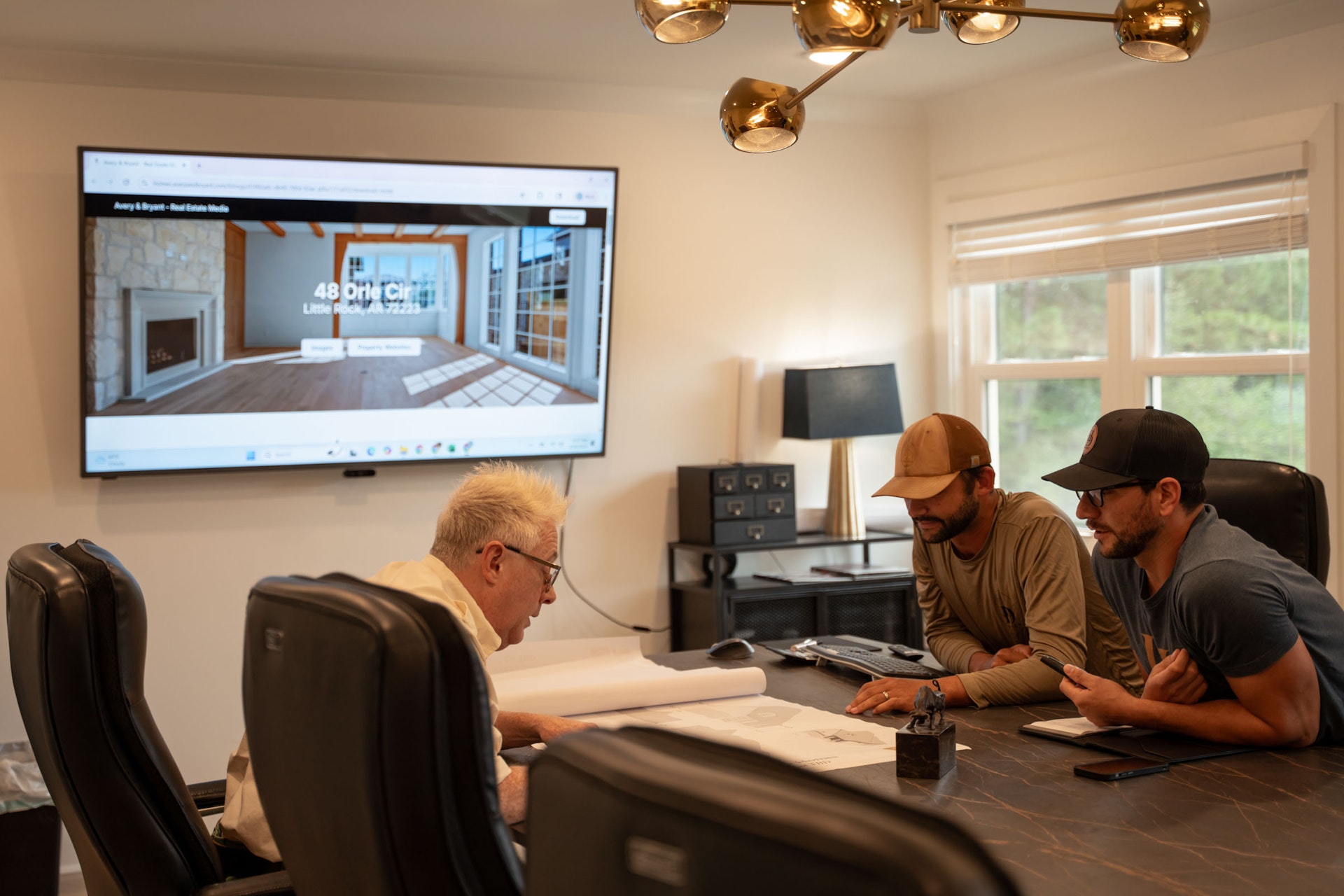 Three men sit at a conference table with papers, discussing plans. A large screen behind them displays a website with a modern home interior. Sunlight streams through windows, and a lamp hangs above the table.